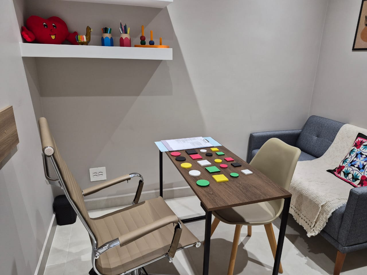Materiais de psicopedagogia sobre a mesa do consultório, com poltrona e sofá para acolhimento